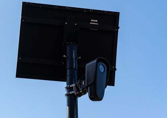 A Flock Safety automated license plate reader camera mounted on a black pole beneath a solar panel in downtown Los Angeles, California, photographed on August 10, 2026. The rear of the solar panel and the camera's mounting brackets, hose clamps and wiring are visible from below against a clear blue sky. The upper floors of a light-colored building bearing painted address numerals stand at lower left, near the corner of West 7th Street and South Alvarado Street in the Westlake neighborhood of Los Angeles, California